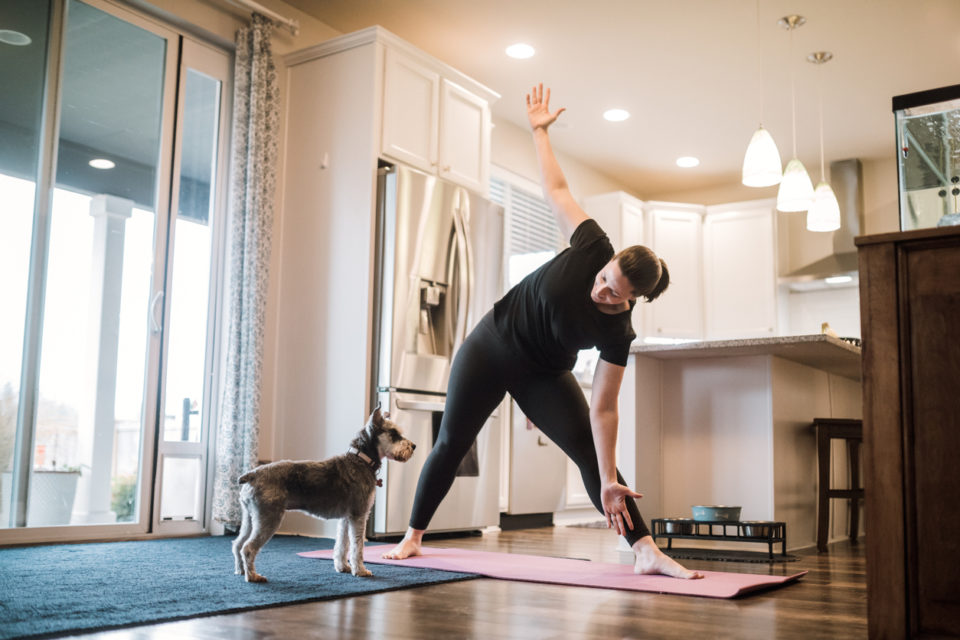 An mature adult woman does yoga and strength training exercises on a mat in her living room, her pet terrier dog keeping her company and trying to play.