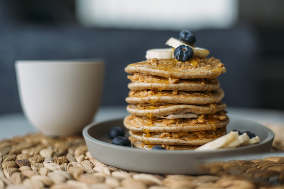 Homemade Pancakes on a grey plate on a woven wicker placemat. There are banana slices and blueberries on top of the panckaes and a white mug in the background.