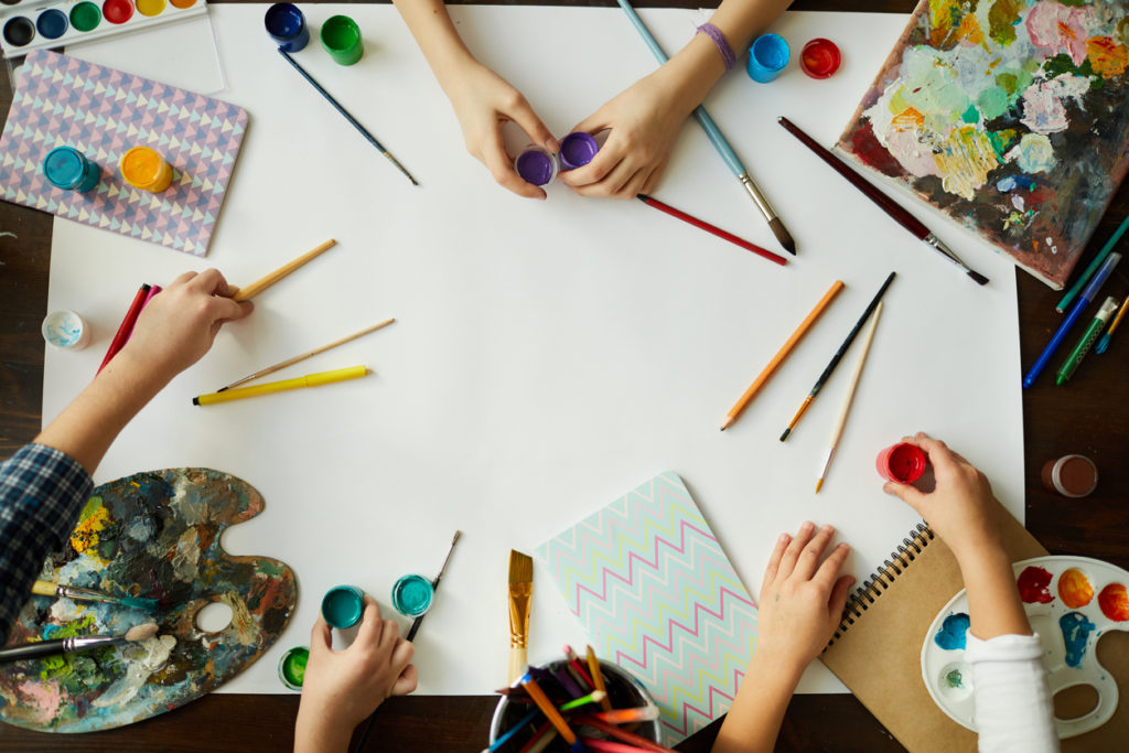 Top view of children's hands over background with art supplies, paints and brushes over white paper